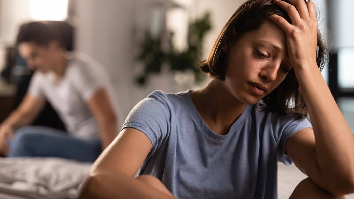 Kvinne som sitter i forgrunnen med hånden på hodet. Mann i bakgrunnen som titter ned.